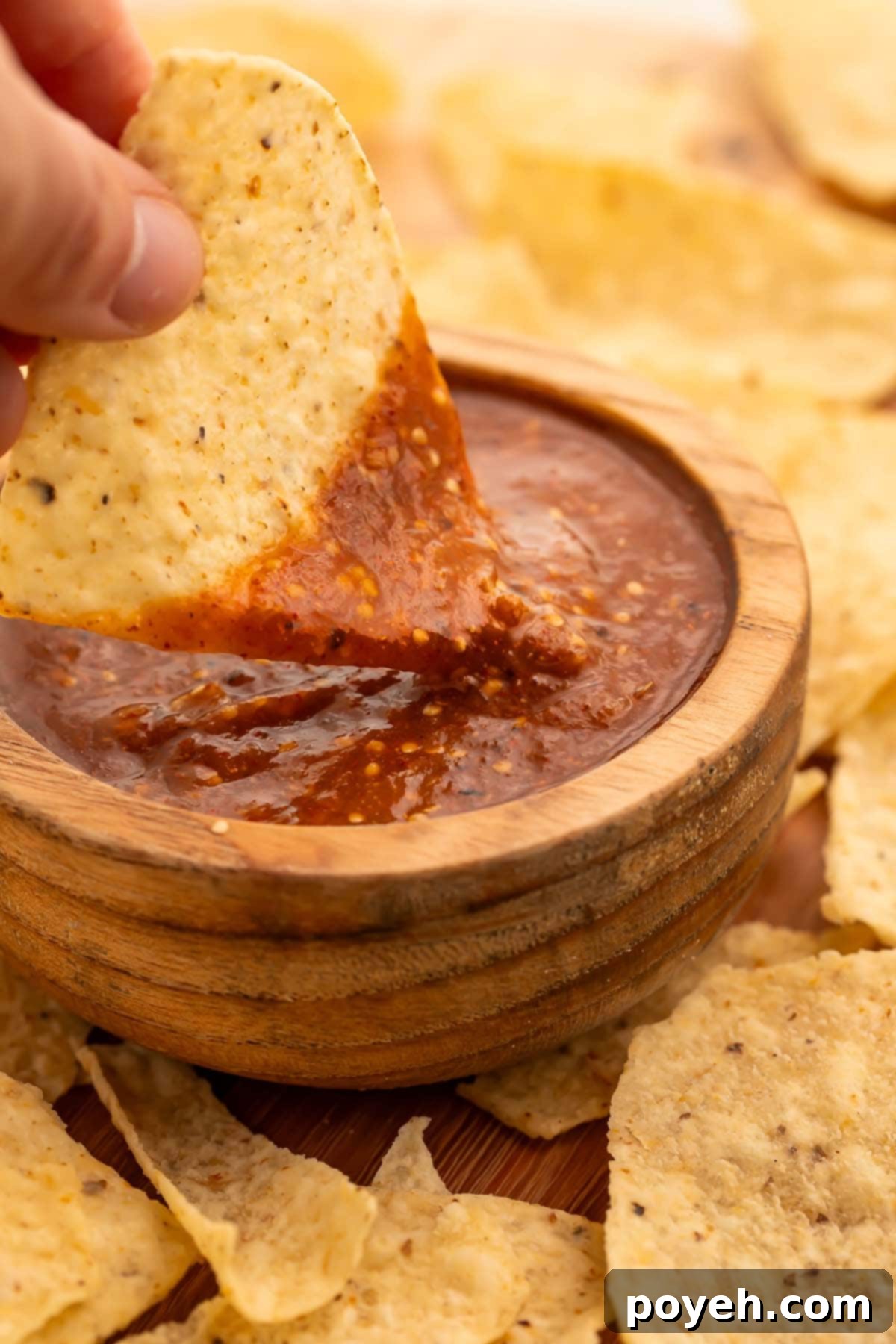A woman's hand holding a tortilla chip with a corner dipped into a deep blood red tomatillo red chili salsa in a bowl.